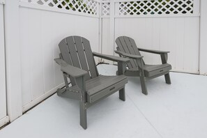 Two Adirondack chairs with metal tables, positioned against a white wooden fence with lattice design.