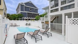 A backyard with a swimming pool, lounge chairs, a white fence, and a two-story house.