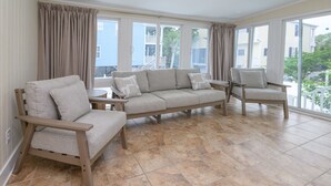 A room with a beige sectional sofa, two wooden chairs, a small table, and large windows with curtains.