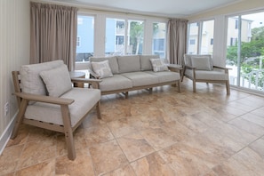 A room with a beige sectional sofa, two wooden chairs, a small table, and large windows with curtains.