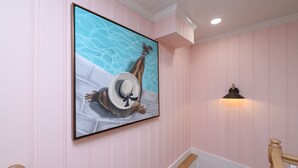 A hallway with pink walls, a framed artwork of a person in a hat by a pool, and a wall-mounted light fixture.