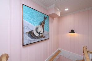 A hallway with pink walls, a framed artwork of a person in a hat by a pool, and a wall-mounted light fixture.