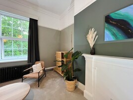 Una habitación con un ventanal grande, una silla, una mesa, una planta y un cuadro en la pared.