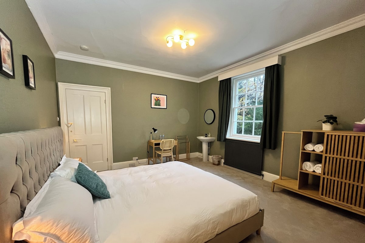 A bedroom with a bed, a desk, a chair, a window, a radiator, and a shelf with towels.