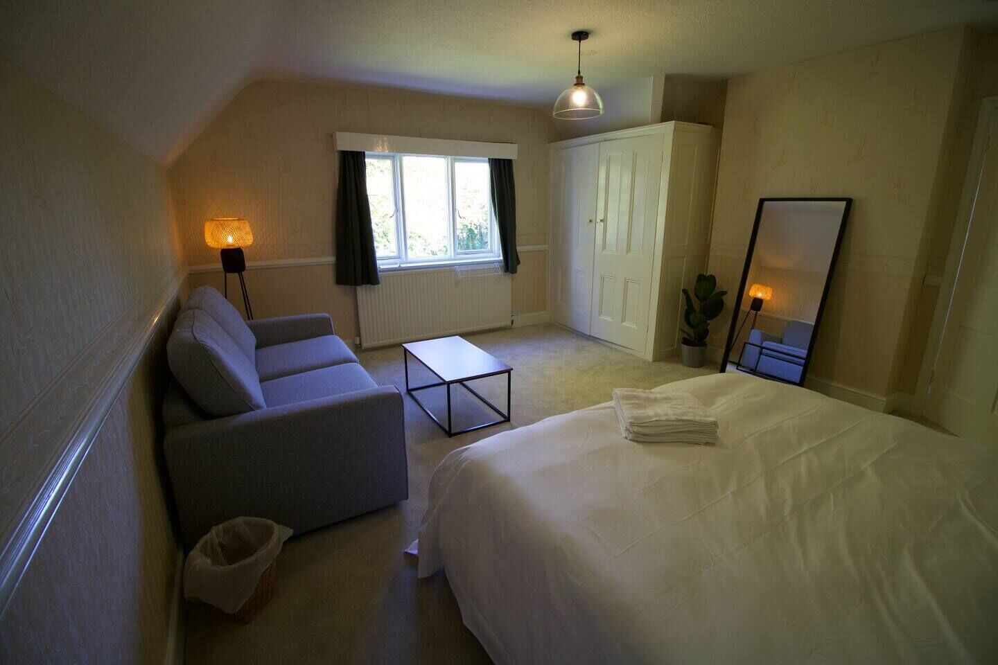 A compact hotel room with a bed, a sofa, a small table, a wardrobe, a window with curtains, and a hanging light fixture.