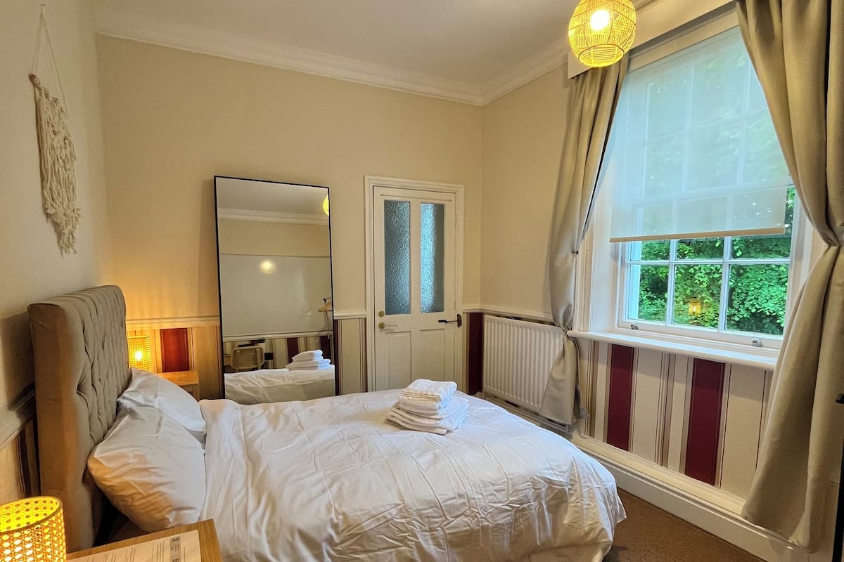 A hotel room with a large bed, a bedside table with a lamp, a mirror, a window with curtains, and a radiator.