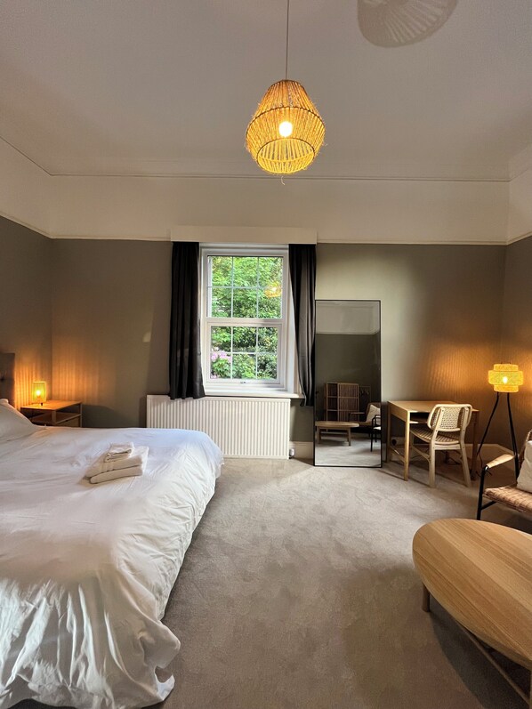 A bedroom with a large bed, a desk, a chair, a standing lamp, and a window with curtains.