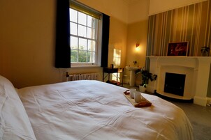 A hotel room with a large bed, a window with curtains, a fireplace, a bedside table with a lamp, and a wall-mounted shelf with decorative items.