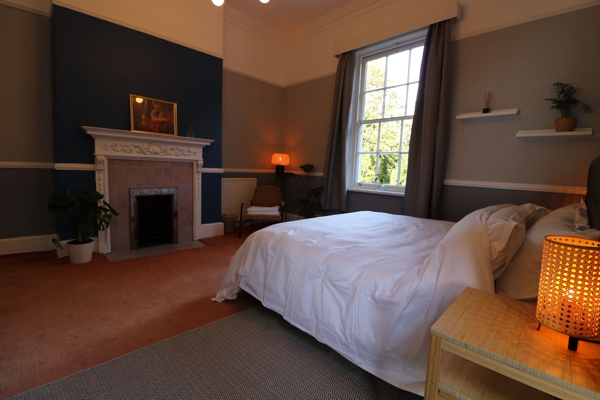 A bedroom with a bed, a fireplace, a window with curtains, a bedside table with a lamp, and a potted plant.