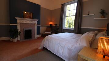 A bedroom with a bed, a fireplace, a window with curtains, a bedside table with a lamp, and a potted plant.