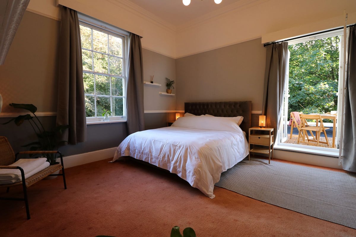 A bedroom with a large bed, a window with curtains, a bedside table with a lamp, and a view of the outdoors.