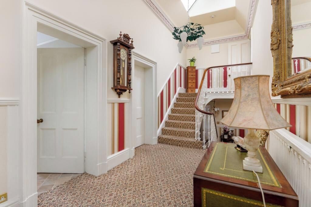 A grand hallway with a patterned carpet, a wooden floor, a staircase with red and white railings, a large mirror, and a table with a lamp.