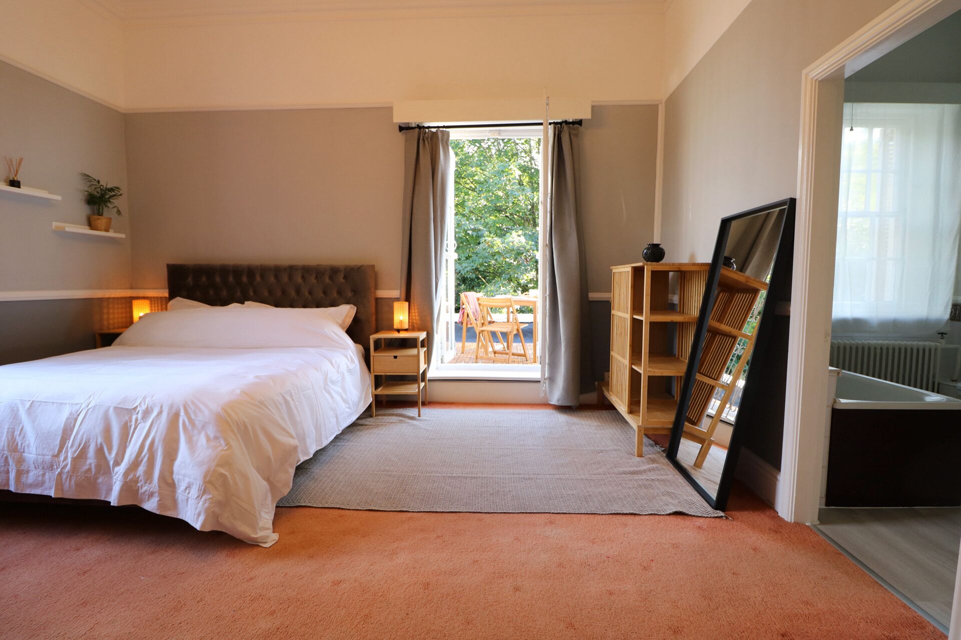 A bedroom with a bed, bedside table, lamp, and a view of the outdoors through a window.