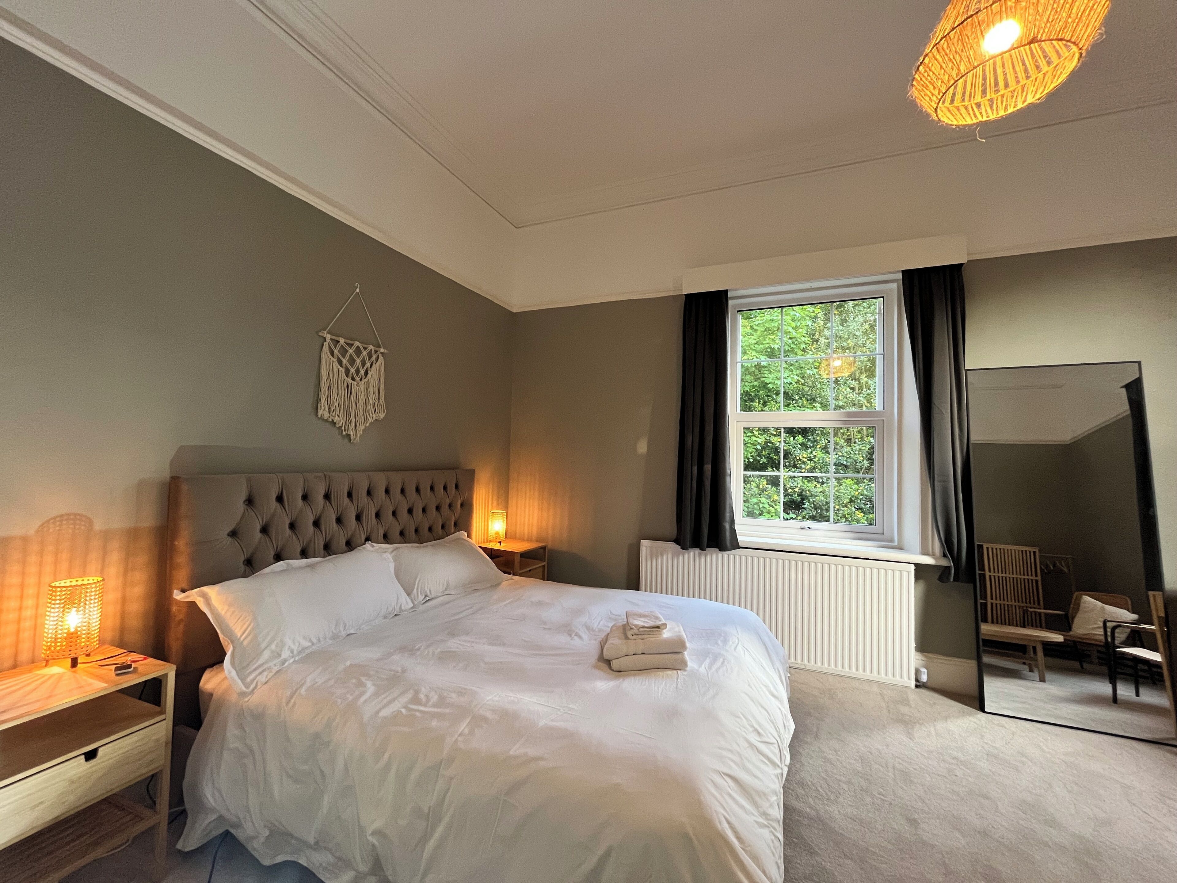 A bedroom with a large bed, bedside lamps, a window with curtains, a mirror, and a macrame wall hanging.