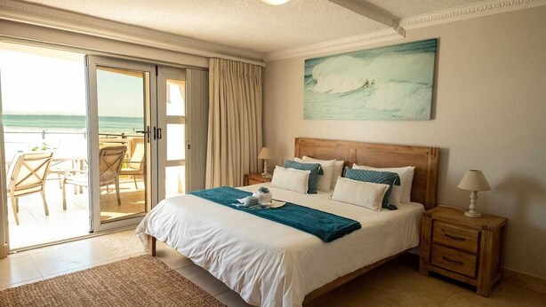 A bedroom with a large bed, a wooden headboard, a painting of a wave, a sliding glass door to a balcony, and a small bedside table. - Stunning ocean vistas, near top surf spots, shops; private, all amenities. (Jeffreys Bay)