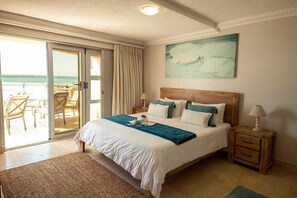 Une chambre avec un grand lit, une tête de lit en bois, un tableau représentant une vague, une porte-fenêtre coulissante donnant sur un balcon et une petite table de chevet.