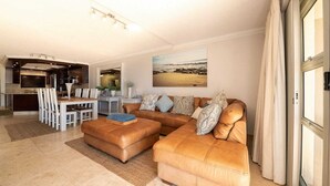 A modern living room with a brown leather sofa, a dining table with chairs, and a kitchen in the background. - Stunning ocean vistas, near top surf spots, shops; private, all amenities. (Jeffreys Bay)