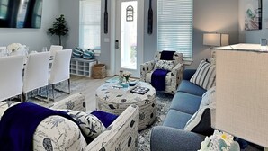 A modern living room with a dining area, featuring a ceiling fan, large windows with blinds, and a TV mounted on the wall. - The Blue Pearls - Luxe Home, Close to Beach + Gulf Views! (Panama City Beach)