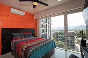 Een slaapkamer met een groot bed, een plafondventilator en uitzicht op een stadsgezicht door een groot raam.