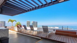 Una terraza en la azotea con vistas al océano, que incluye una cubierta de madera, sillas de metal y una pequeña mesa.