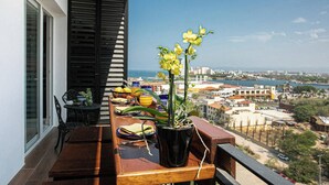 Een balkon met een houten eettafel, een bloempot met gele bloemen en een stadsgezicht op de achtergrond.
