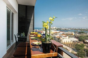 Een balkon met een houten eettafel, een bloempot met gele bloemen en een stadsgezicht op de achtergrond.