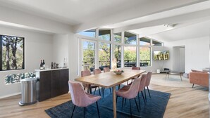 Un comedor amplio con una gran mesa de madera y sillas rosadas, una zona de cocina con encimera oscura, y ventanas grandes que ofrecen vistas a árboles y una terraza.