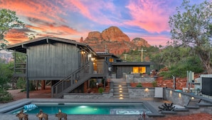 Una casa moderna con piscina, iluminación exterior y vistas a formaciones rocosas rojizas.