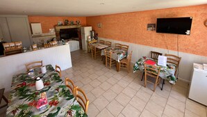 Een eethoek met een keuken op de achtergrond, houten stoelen, een tafel met een bloemendecoratie en een aan de muur bevestigde televisie.