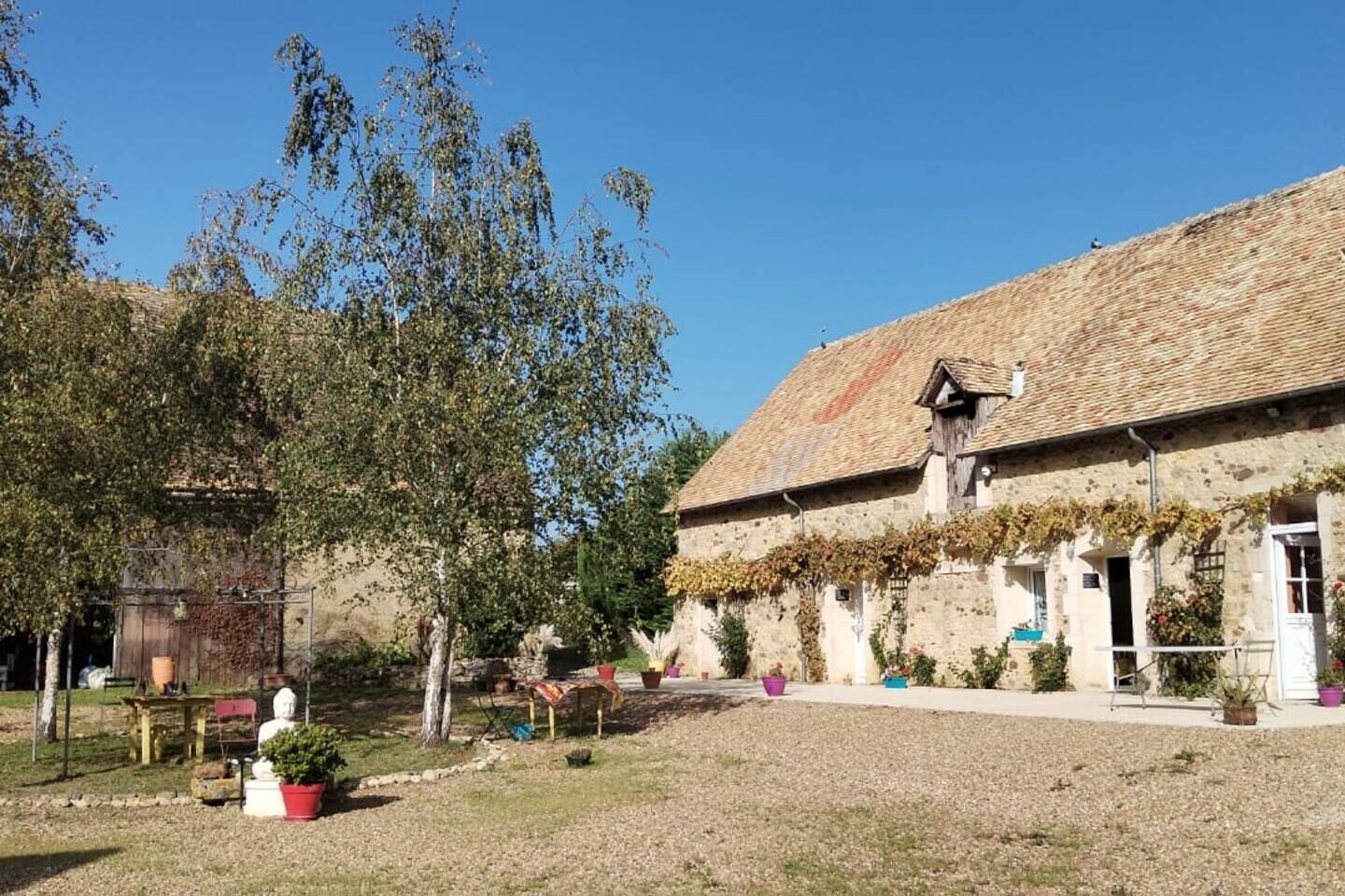 A rustic stone building with a tiled roof, surrounded by a garden with potted plants and a tree.