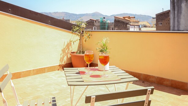 Una terraza en la azotea con una mesa dispuesta para dos personas, dos vasos de una bebida y una planta en maceta.