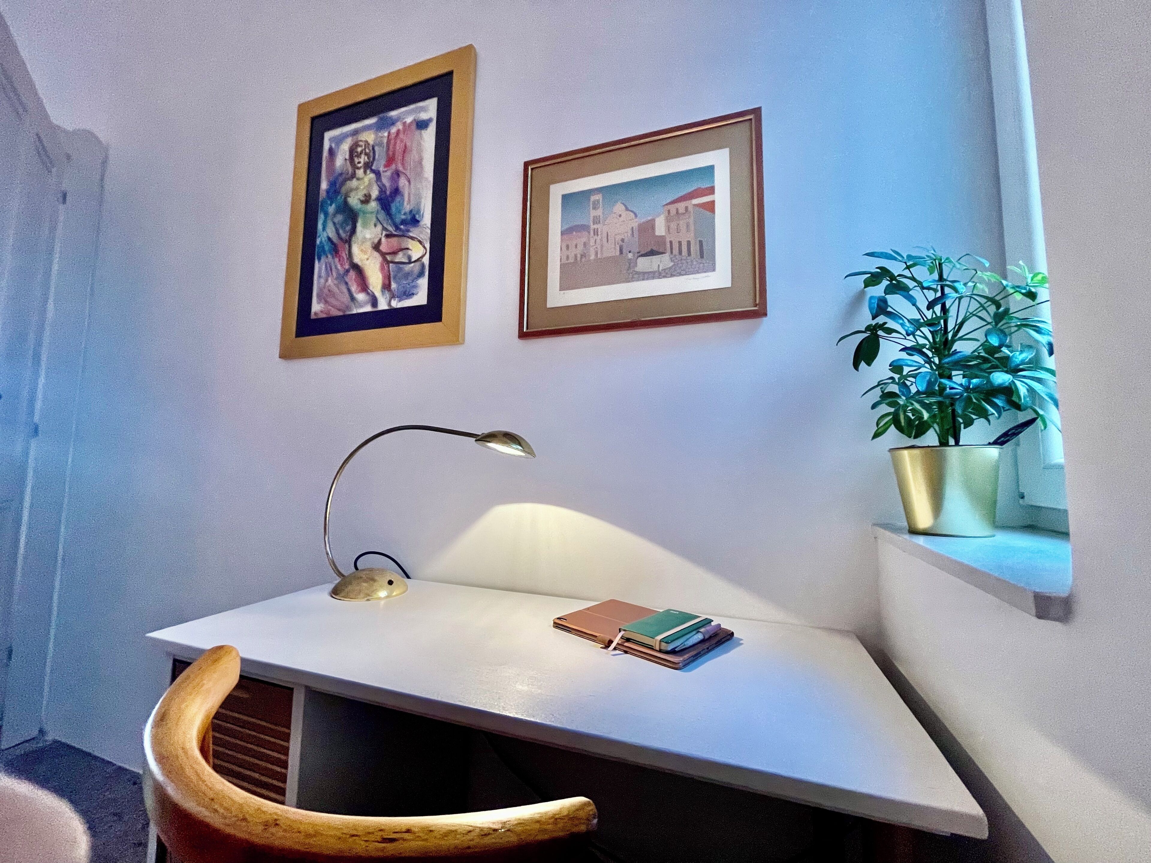A small workspace with a desk, chair, lamp, framed artwork, and a potted plant.