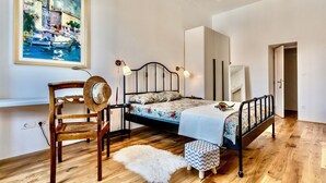 Un dormitorio con una cama, una silla, un pequeño reposapiés, un cuadro y un espejo.