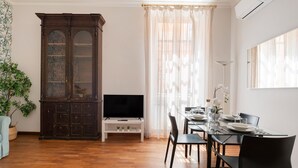Une pièce dotée d’un plancher en bois, d’un buffet en bois foncé avec des portes en verre, d’un téléviseur à écran plat posé sur un meuble blanc, d’une table à manger avec des chaises, et d’une fenêtre ornée de rideaux légers.