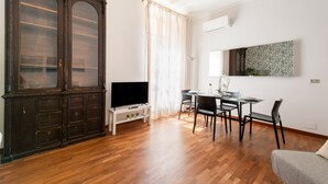 Un salon comprenant un grand meuble en bois foncé, un téléviseur à écran plat posé sur un meuble blanc, une table à manger avec des chaises et une grande fenêtre ornée de rideaux légers.