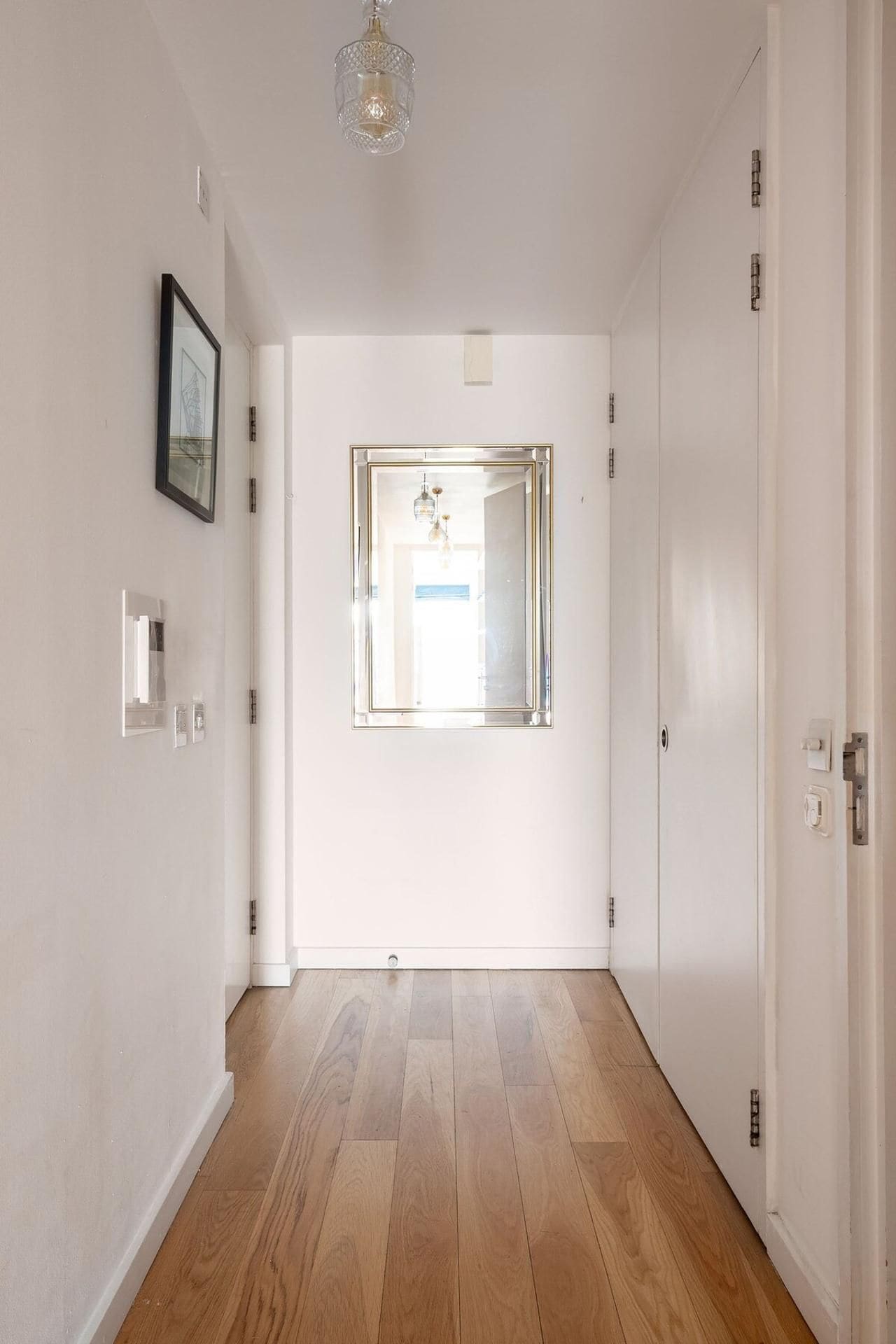 Un pasillo con suelo de madera, paredes blancas, un cuadro enmarcado, un accesorio de iluminación y una puerta con espejo.