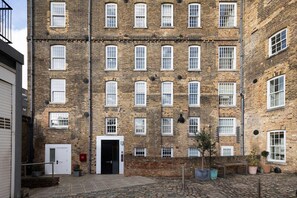 A multi-story brick building with a central entrance, multiple windows, and a paved courtyard with potted plants.