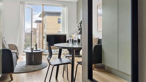Un salon moderne avec une table ronde, deux chaises, un canapé et une grande fenêtre donnant sur des immeubles.