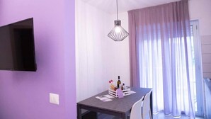 Une pièce avec un mur violet, un téléviseur à écran plat fixé au mur, une table à manger avec des chaises, un luminaire suspendu et une fenêtre avec des rideaux.