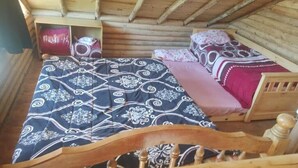 Puinen mökki, jonka sisustuksessa on sänky, jossa on kuviollinen päiväpeite ja tyynyjä, puinen pääty sekä pieni hylly, jossa on esineitä.