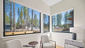 A modern room with large windows, a wooden floor, a grey armchair, a small round table, and a bedside table with a lamp. - Modern Caldera Home with Access to Forest House (Bend)