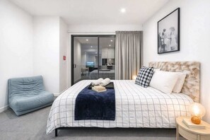 Moderni hotellihuone, jossa on suuri sänky, oleskelualue, televisio ja ruokailutila, joka on nähtävissä lasiovesta.