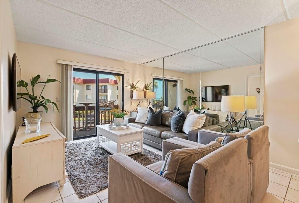 A modern living room with a sofa, coffee table, and a large mirror. There is a balcony with a view of buildings.