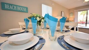 A dining table set with white plates, blue napkins, and clear glassware, with a 'BEACH HOUSE' sign on the wall.