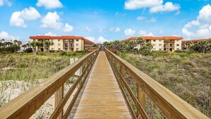 A wooden boardwalk leading to a hotel with red roofs, surrounded by grass and palm trees.