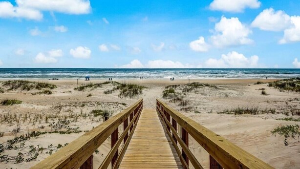 A wooden boardwalk leading to a beach with dunes and a clear blue sky.