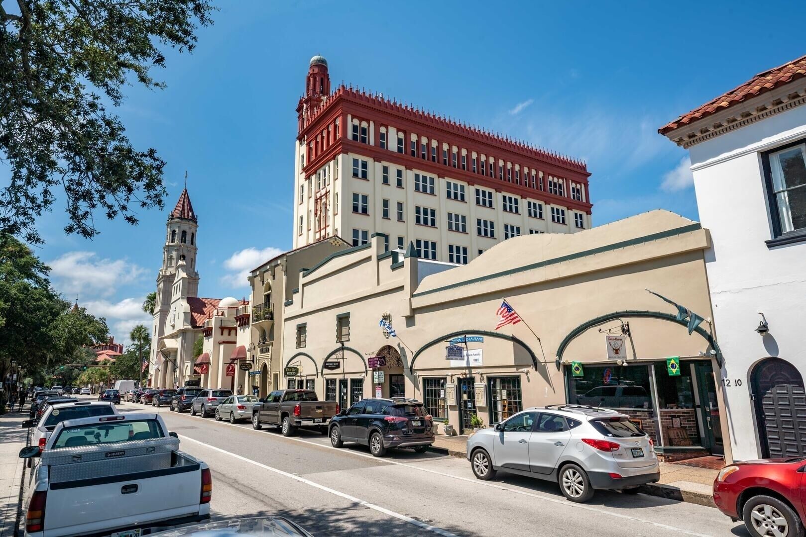 A street view with a prominent building featuring a clock tower, multiple storefronts, and parked cars.