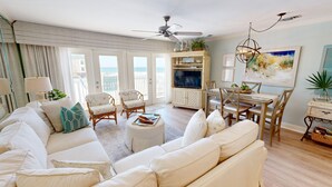A spacious living room with a large sofa, two armchairs, a dining table with chairs, a ceiling fan, and a TV.