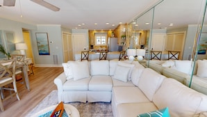 A spacious living room with a large white sectional sofa, a dining area with wooden chairs, and a kitchen with wooden cabinets and a central island.