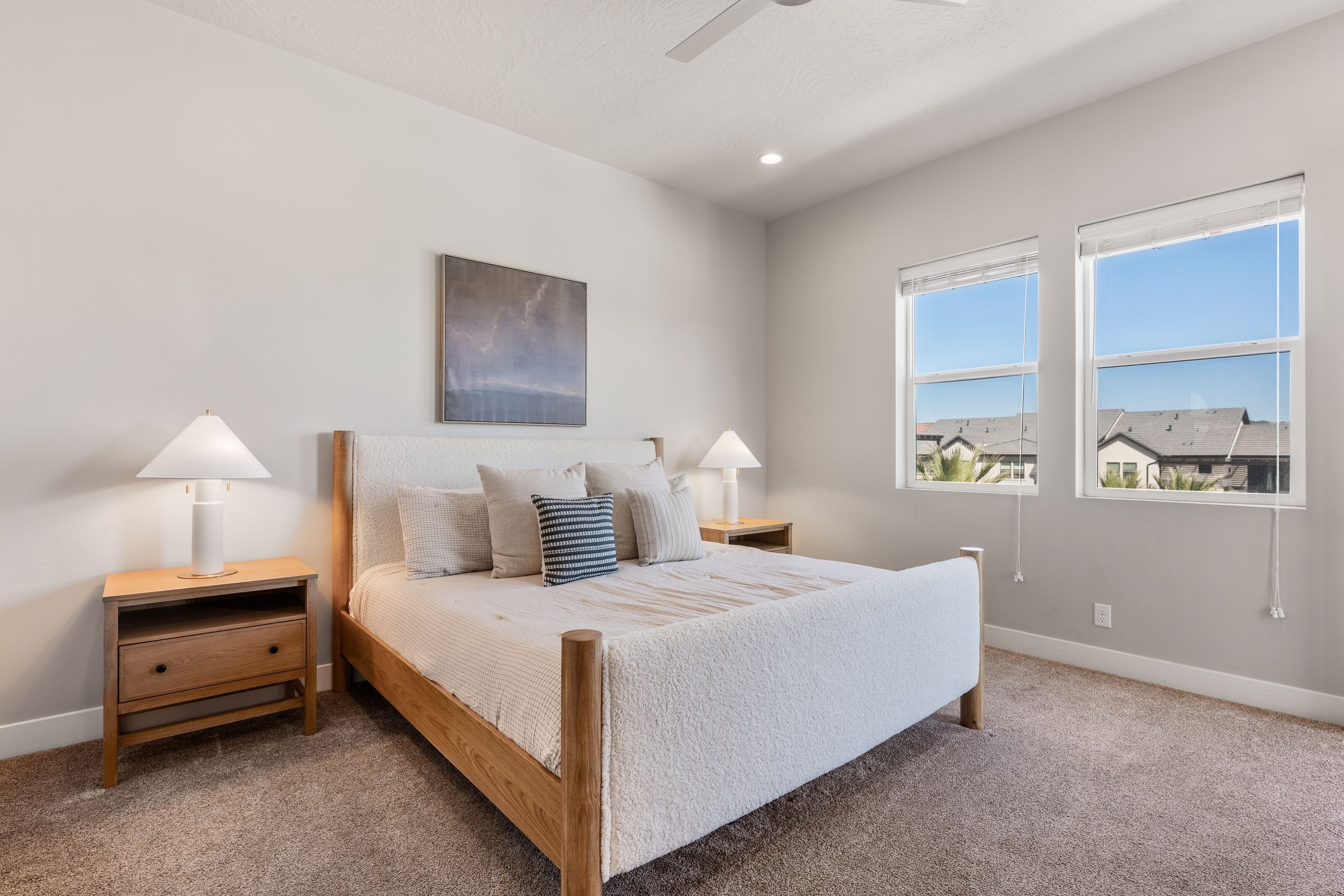 A bedroom with a wooden bed, two bedside tables with lamps, a large window with blinds, and a painting on the wall.